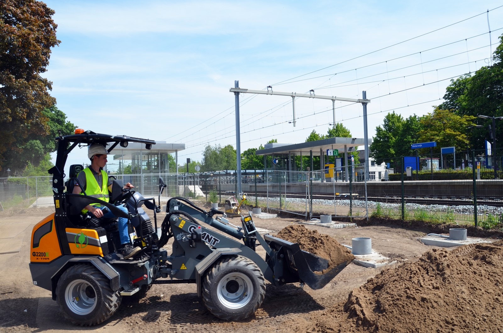 Ouvrier sur un chantier de terrassement autour d'une gare avec la CHARGEUSE électrique GIANT G2200E X-TRA et son godet pour charger et transporter de la terre en toute sécurité.